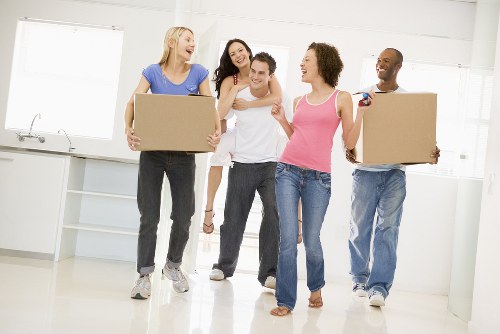 A group of five diverse people, including women and a man, standing in a bright, modern kitchen with a minimalist design, white walls, large windows, and white cabinetry. Two women are holding medium-sized cardboard boxes, ready for a house move, while the other three are smiling and engaging in conversation. The individuals are dressed casually in jeans and t-shirts, with some wearing sandals or sneakers. The scene captures a cheerful and organized atmosphere typical of a professional house removal service, with no visible furniture or appliances, emphasizing the focus on packing and moving personal belongings. The natural light streaming in from the windows highlights the clean, neutral colours of the space, subtly suggesting a residential setting in an urban area, possibly in the vicinity of London, where Mayfair Man with Van operates. The overall scene reflects the effectiveness and friendly approach of local removals expertise in a comfortable home environment, aligned with services offered by Mayfair Man with Van.
