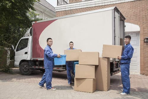 Three male movers dressed in blue work uniforms are loading large cardboard boxes onto a white and maroon removals truck parked on a paved driveway. The boxes vary in size, with some stacked on the ground and others being held by the movers, who appear engaged in carrying or positioning the items. The scene takes place in a residential area, with a brick house visible on the right and greenery in the background, suggesting a suburban environment. The lighting indicates daylight, and the overall setting reflects a house removal operation typical of Mayfair, London, with Mayfair Man with Van providing professional moving services. The trucks and boxes are arranged systematically, emphasizing organized packing and transportation of household belongings.