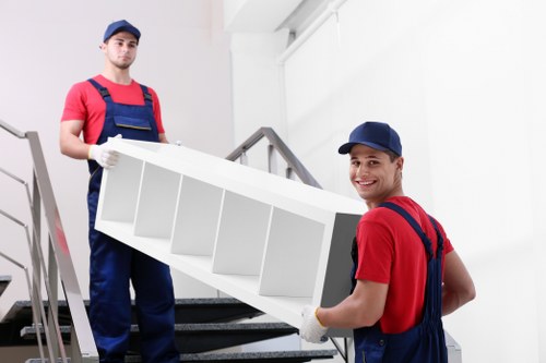 Two male movers, dressed in red shirts, dark blue overalls, and matching navy caps, are carrying a white, multi-compartment shelf or storage unit down a set of indoor stairs. One mover, positioned in the foreground on the right, is holding the shelf with both hands, smiling towards the camera, while the other, standing behind on the left, supports the opposite side with one hand and looks ahead. The shelf has a smooth, matte finish and is divided into several open cubby-style sections. The stairway features a metal handrail on the left side, and the background shows a bright, clean interior with white walls and natural lighting. This scene is set inside a residential property in Mayfair, London, and is part of a professional house removal service, as indicated by the clean, organized environment and the movers' uniforms, reflecting the offerings of Mayfair Man with Van's removals expertise.