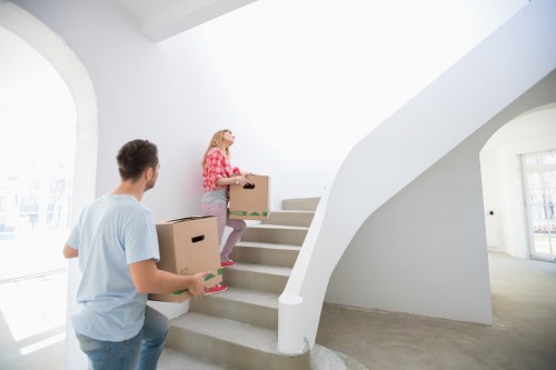In a brightly lit, modern residential interior with white walls and large windows allowing natural light, a man and a woman are engaged in house moving activities as part of a removals service. The man, dressed casually in a light blue T-shirt, is seen from the back holding a medium-sized cardboard box, preparing to carry it upstairs. The woman, wearing a red and white checked shirt and pink trousers, is walking up a curved staircase with a wooden handrail, carrying a similar cardboard box. The staircase has white painted walls and wooden steps, with additional cardboard boxes stacked behind the woman on the stairs. In the background, a spacious, airy room can be seen featuring a minimalist design, with bright natural daylight streaming through large arched windows, suggesting a house located within a residential area in or near Mayfair, central London. The scene captures a professional removal effort consistent with house relocation services offered by Mayfair Man with Van, highlighting the careful handling of household items during the move.