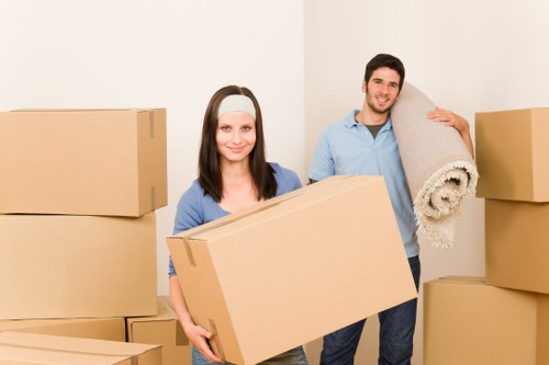 A woman and a man are standing inside a room during a house move, surrounded by large cardboard boxes of various sizes, some stacked against the plain white wall. The woman, with shoulder-length dark hair and a white headband, is holding a medium-sized cardboard box close to her chest, smiling at the camera. She is dressed in a casual blue top. The man, with short dark hair, is standing behind her, carrying a rolled-up beige carpet or rug over his shoulder, also smiling. The background shows a plain white wall, and the scene is well-lit, suggesting natural or studio lighting. The setting appears to be an interior space preparing for a move, with minimal furniture visible, emphasizing the packing activity. The image reflects a professional house removal service environment, consistent with the offerings of Mayfair Man with Van, a provider specializing in local removals in London, close to the postcode area of Mayfair.