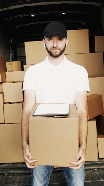 A professional mover from Mayfair Man with Van, wearing a navy blue t-shirt with a blue logo on the chest and dark trousers, is inside a bright, empty room with white walls and a light wooden floor. T