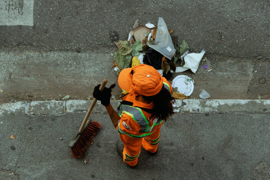 A waste collection worker dressed in high-visibility orange clothing, including a jacket, trousers, and a safety helmet, is standing on a sidewalk next to a pile of mixed household rubbish. The worker