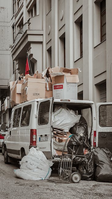 A waste collection worker dressed in high-visibility orange clothing, including a jacket, trousers, and a safety helmet, is standing on a sidewalk next to a pile of mixed household rubbish. The worker